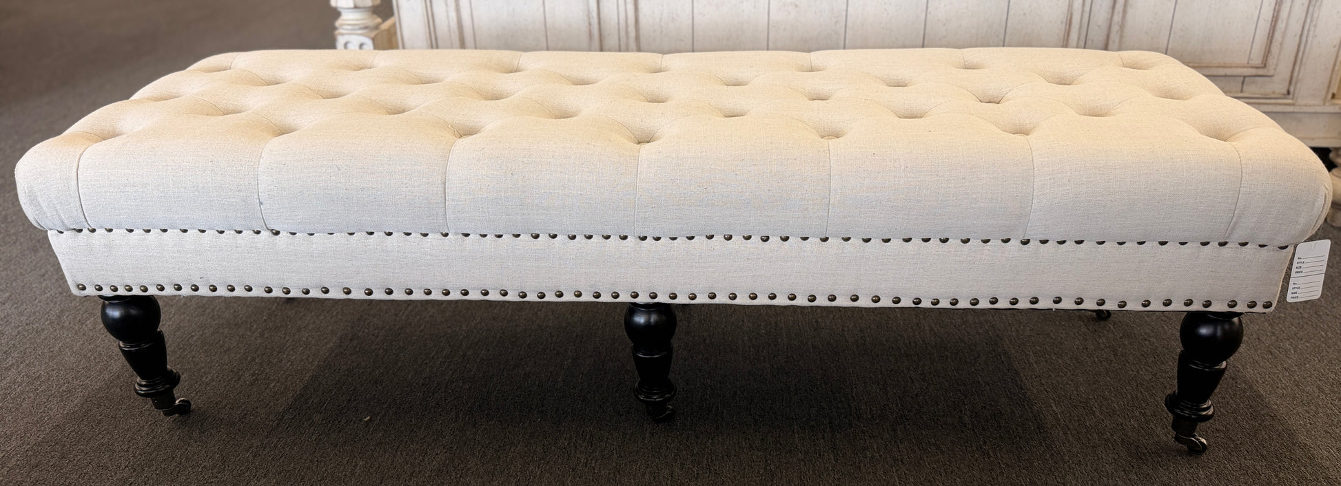 A long rectangular bedroom bench upholstered in cream linen with deep button tufting, antique bronze nailhead trim, and six black turned legs on casters in good condition.