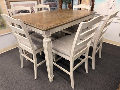 A two-tone counter-height dining set featuring a brown wood top rectangular table and six distressed white chairs with textured grey upholstered seats.