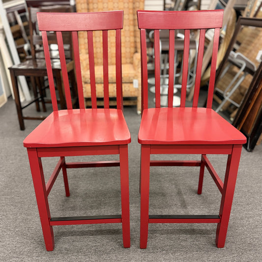 A pair of bright red wooden counter-height barstools featuring vertical slatted backrests, contoured seats, and contrasting black footrest bars.