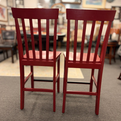 A pair of bright red wooden counter-height barstools featuring vertical slatted backrests, contoured seats, and contrasting black footrest bars.