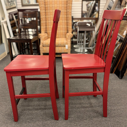 A pair of bright red wooden counter-height barstools featuring vertical slatted backrests, contoured seats, and contrasting black footrest bars.