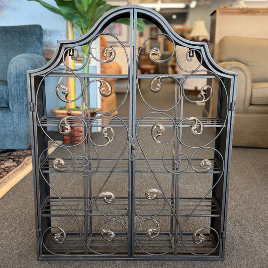 A decorative wall-mounted metal cabinet featuring an arched top, multi-tier interior shelves, and two intricate scrollwork doors that latch in the center.