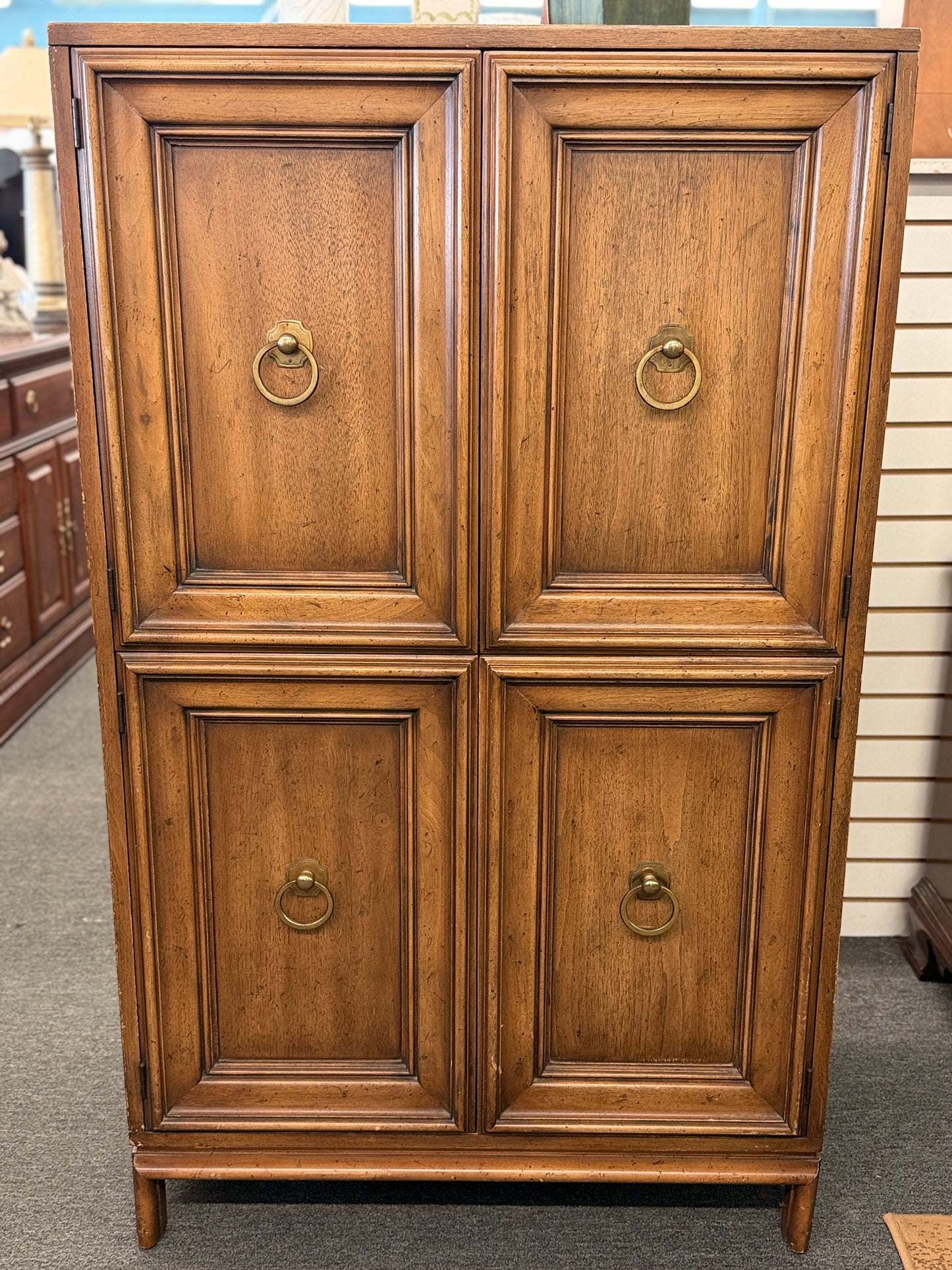 A vintage wooden armoire storage cabinet featuring a symmetric four-panel door facade with recessed molding and antiqued brass ring pull handles.