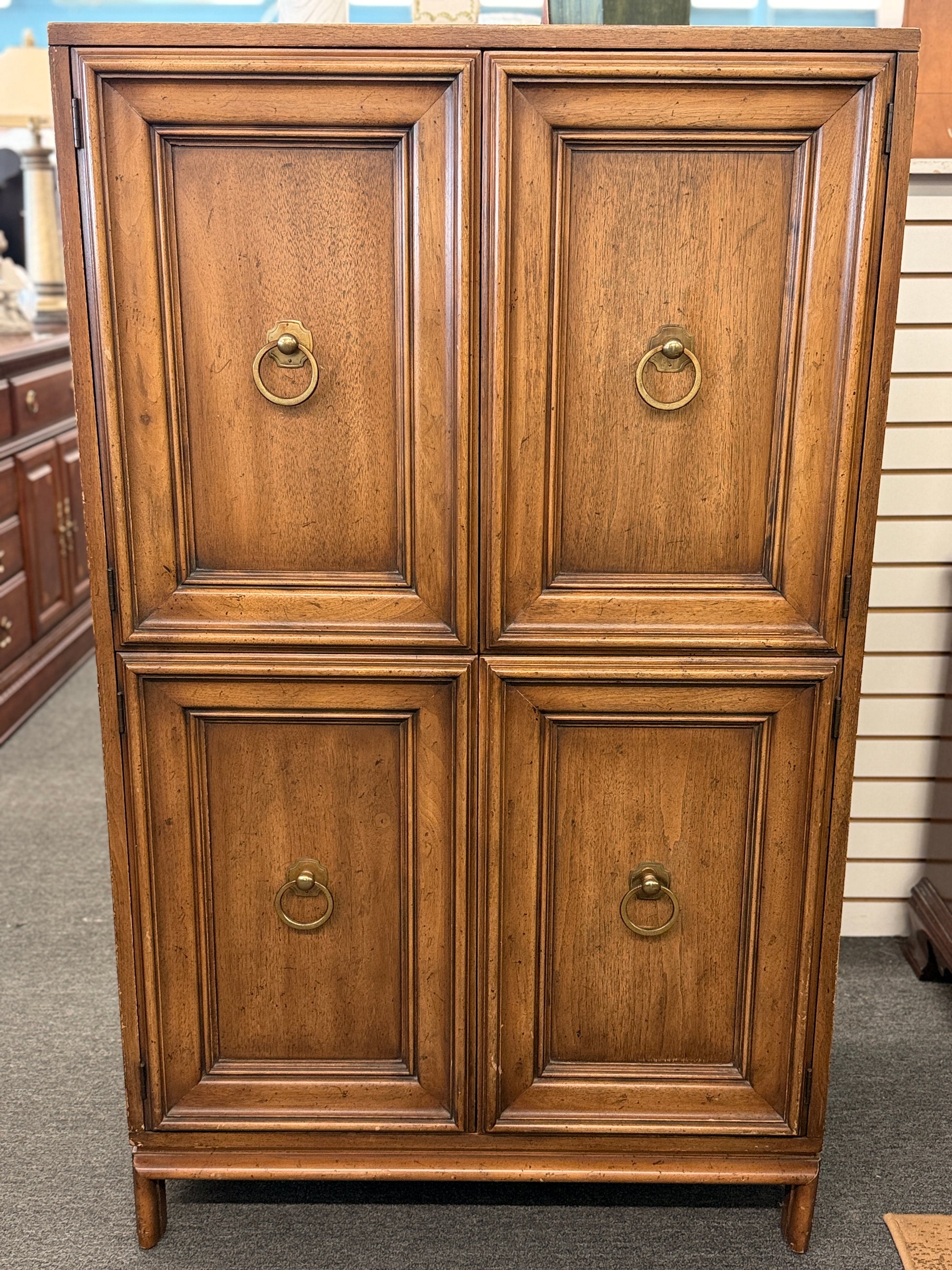 A vintage wooden armoire storage cabinet featuring a symmetric four-panel door facade with recessed molding and antiqued brass ring pull handles.