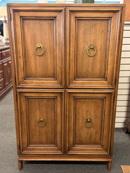 A vintage wooden armoire storage cabinet featuring a symmetric four-panel door facade with recessed molding and antiqued brass ring pull handles.