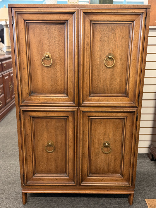 A vintage wooden armoire storage cabinet featuring a symmetric four-panel door facade with recessed molding and antiqued brass ring pull handles.