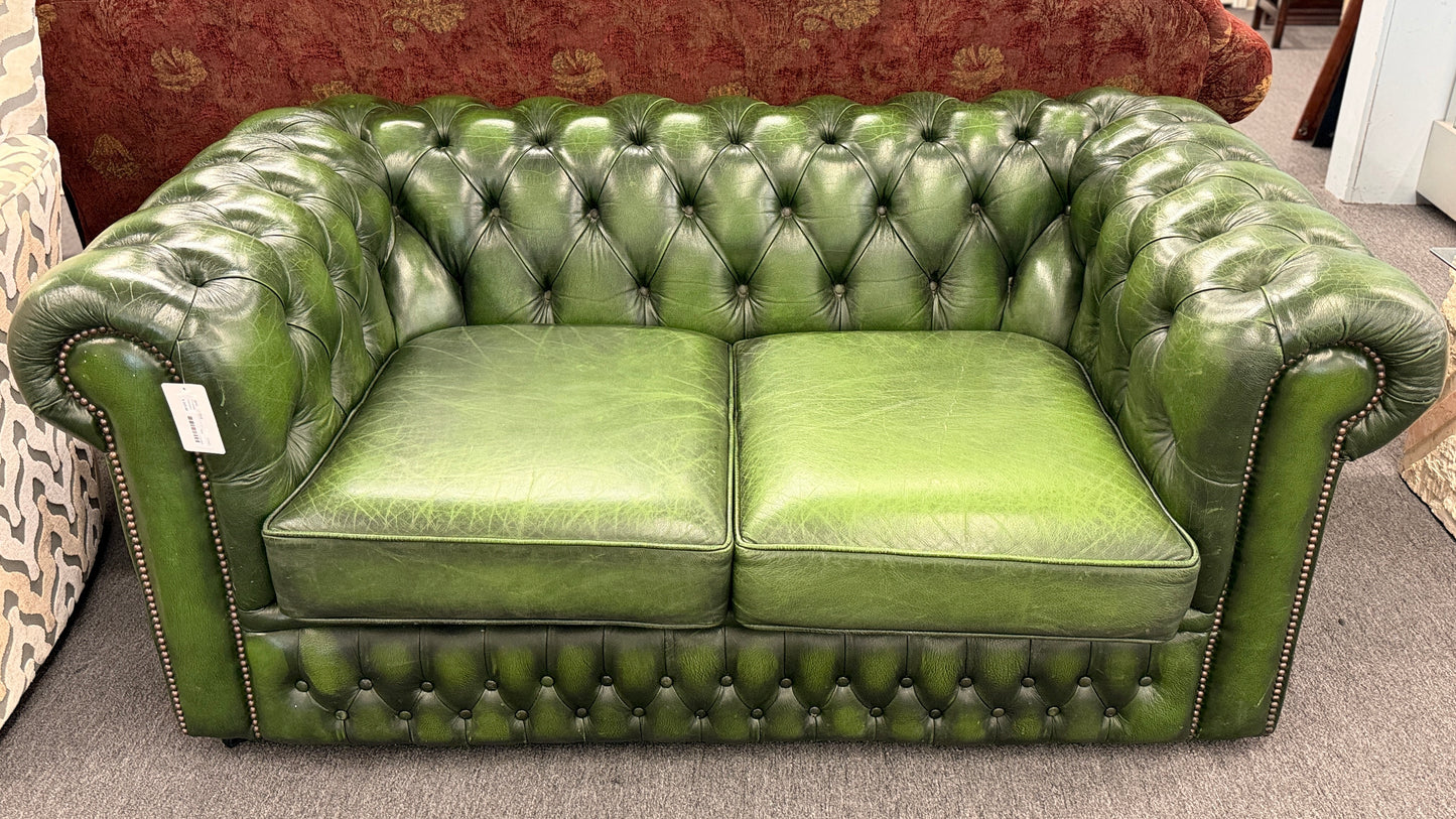 A classic Chesterfield-style green leather two-seater loveseat featuring deep button tufting, rolled arms, and brass nailhead trim on a wooden frame.