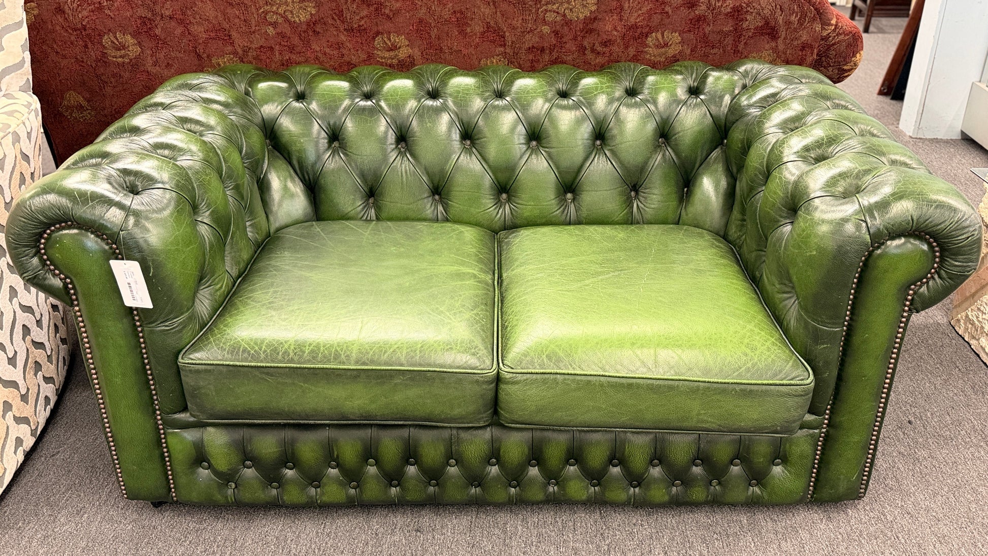 A classic Chesterfield-style green leather two-seater loveseat featuring deep button tufting, rolled arms, and brass nailhead trim on a wooden frame.