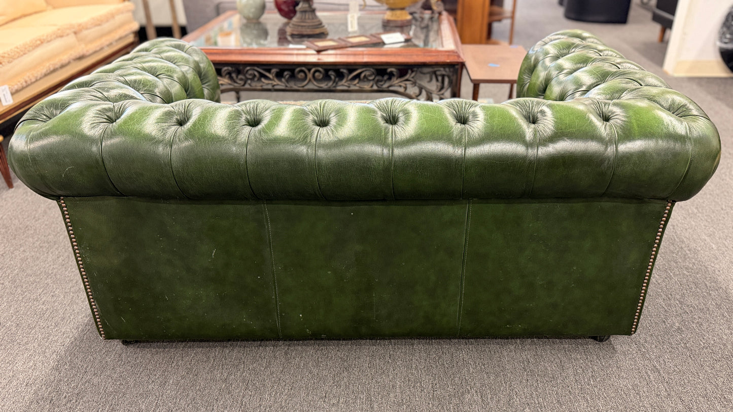 A classic Chesterfield-style green leather two-seater loveseat featuring deep button tufting, rolled arms, and brass nailhead trim on a wooden frame.