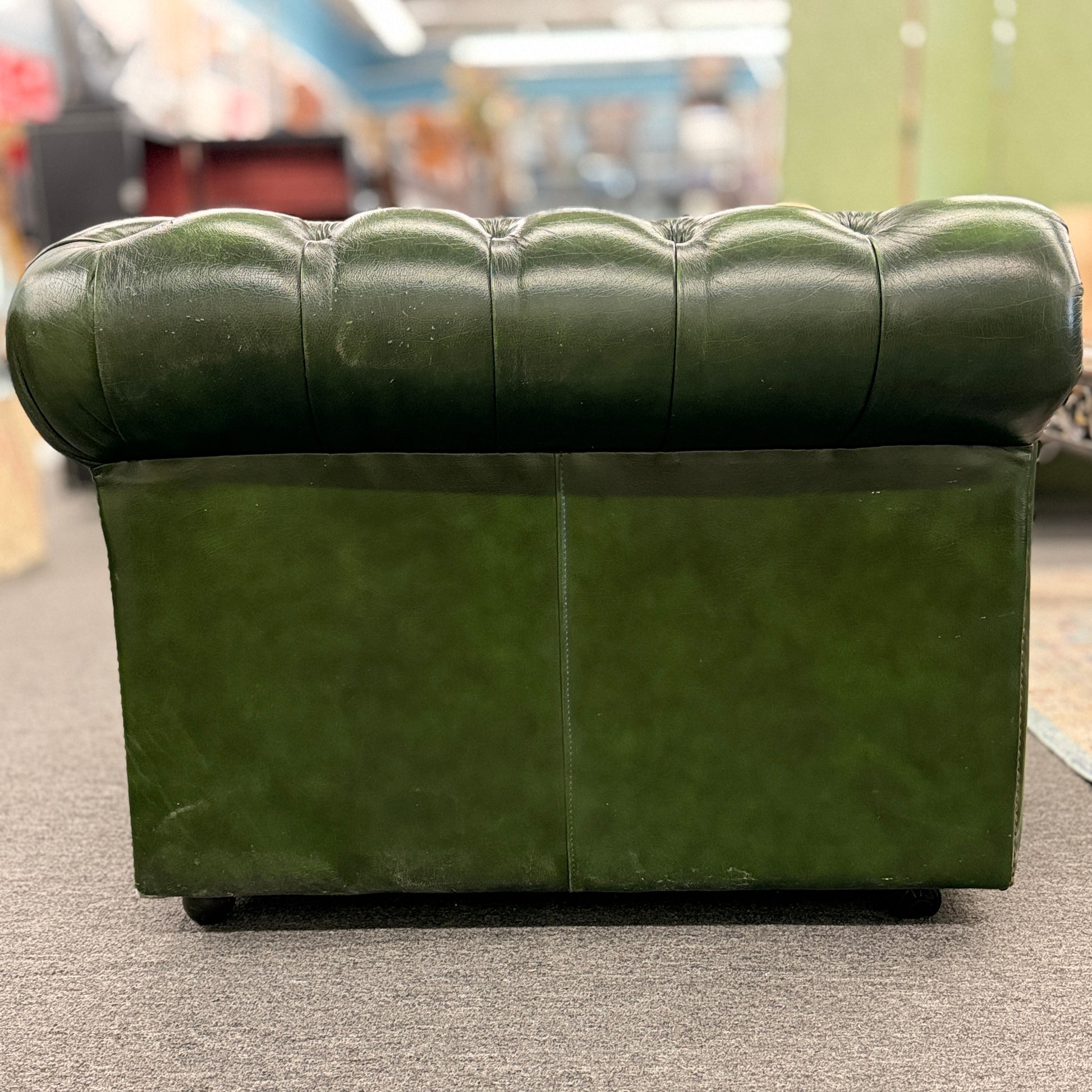 A classic Chesterfield-style green leather two-seater loveseat featuring deep button tufting, rolled arms, and brass nailhead trim on a wooden frame.