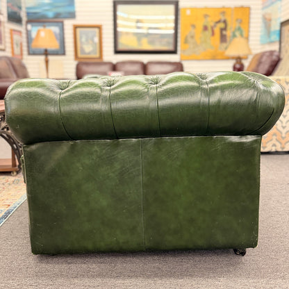 A classic Chesterfield-style green leather two-seater loveseat featuring deep button tufting, rolled arms, and brass nailhead trim on a wooden frame.