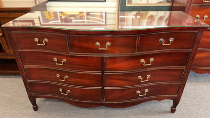 A vintage multi-drawer dresser in a rich reddish-brown wood finish with a serpentine front, brass handles, and a glass top in good condition.