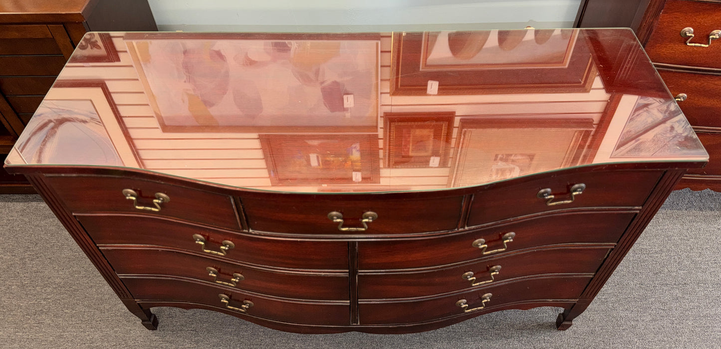 A vintage multi-drawer dresser in a rich reddish-brown wood finish with a serpentine front, brass handles, and a glass top in good condition.