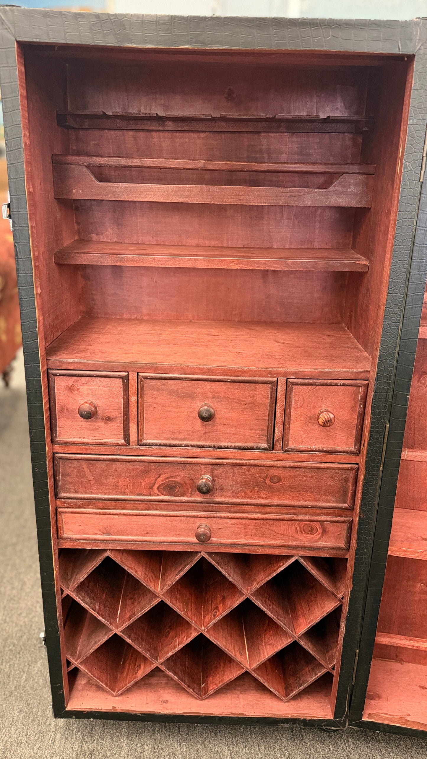 A tall hinged trunk-style bar cabinet featuring interior shelving, small utility drawers, and a lower lattice rack for wine bottle storage.