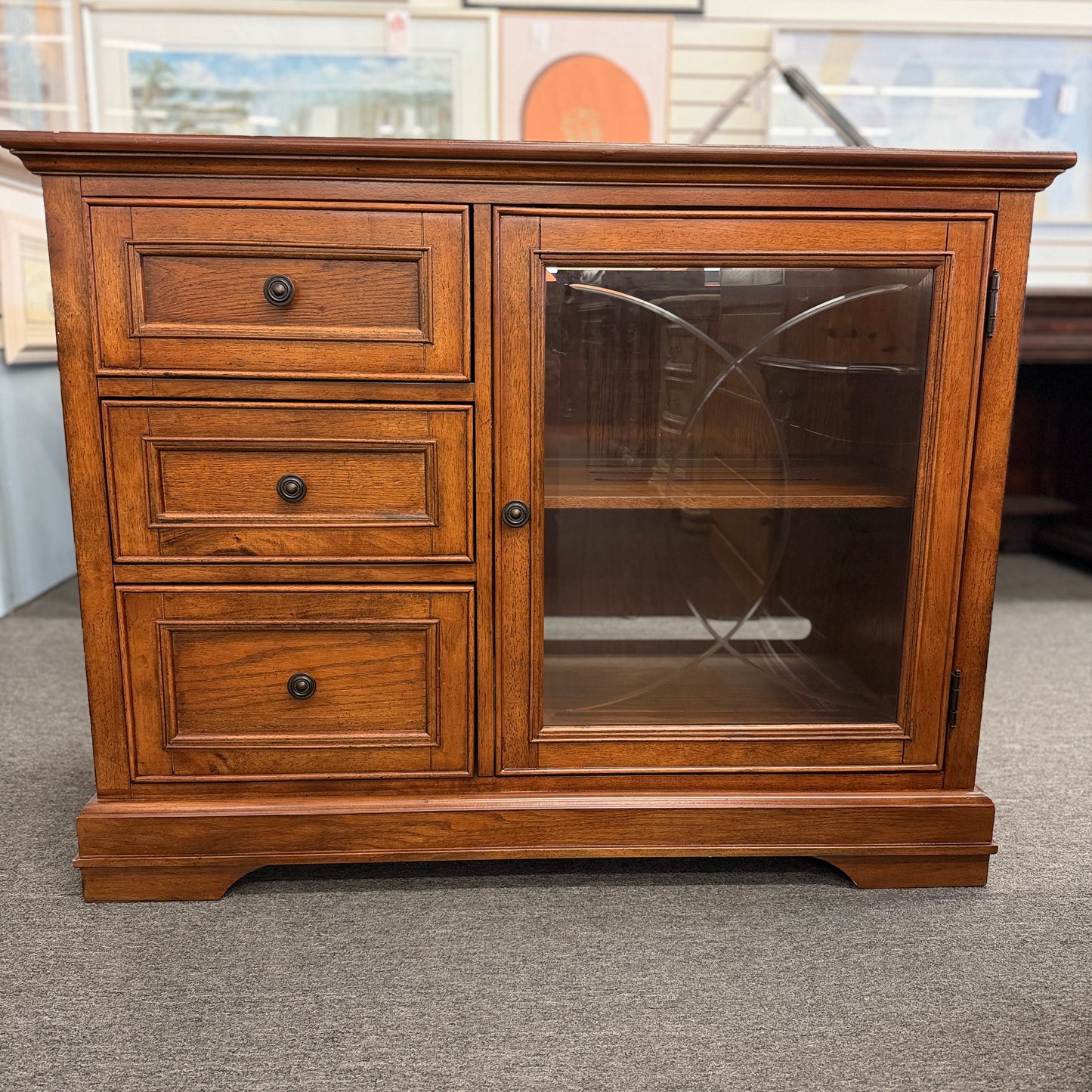 Medium-tone wood storage cabinet featuring three stacked drawers, a glass-paneled cabinet door with interior shelving, and a classic versatile design.