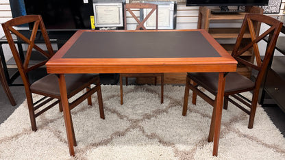 A wooden folding dining table set featuring a dark brown inset playing surface and four matching folding chairs with dark brown upholstered seat cushions.
