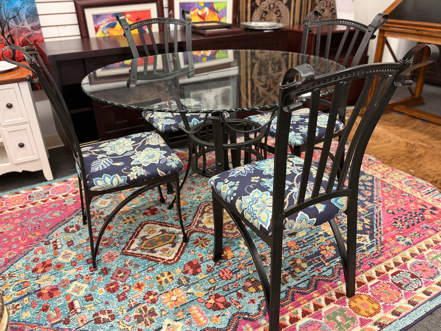 A wrought iron-style dining set featuring a round glass tabletop on a black metal base with four matching black metal chairs with blue and yellow floral fabric seats.