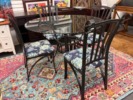 A wrought iron-style dining set featuring a round glass tabletop on a black metal base with four matching black metal chairs with blue and yellow floral fabric seats.