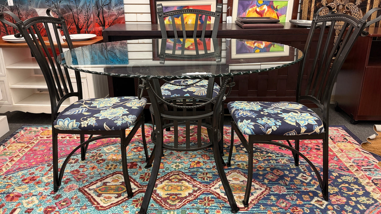 A wrought iron-style dining set featuring a round glass tabletop on a black metal base with four matching black metal chairs with blue and yellow floral fabric seats.