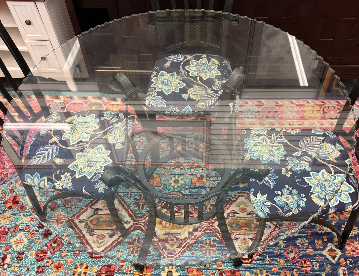 A wrought iron-style dining set featuring a round glass tabletop on a black metal base with four matching black metal chairs with blue and yellow floral fabric seats.