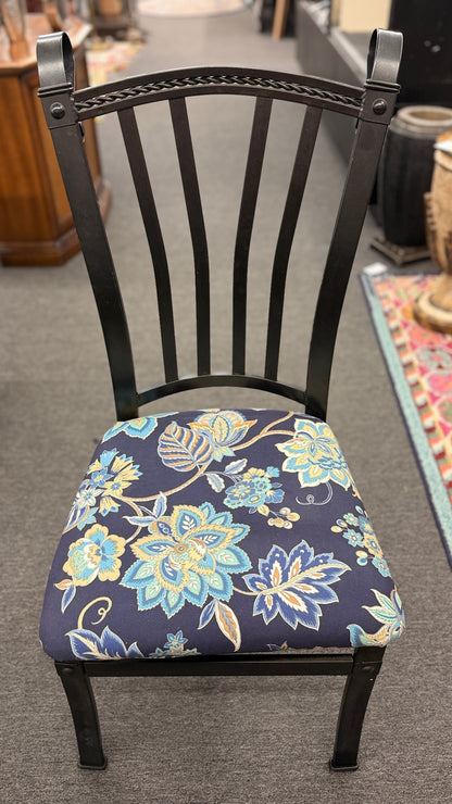 A wrought iron-style dining set featuring a round glass tabletop on a black metal base with four matching black metal chairs with blue and yellow floral fabric seats.