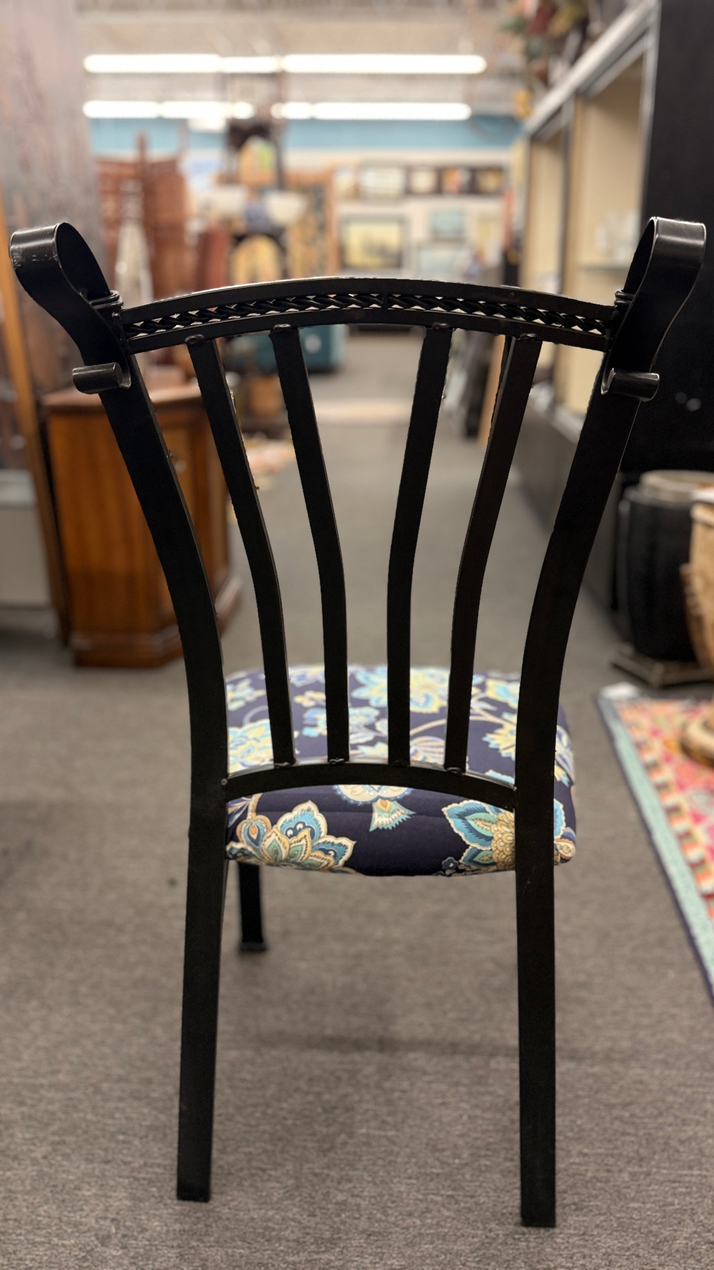 A wrought iron-style dining set featuring a round glass tabletop on a black metal base with four matching black metal chairs with blue and yellow floral fabric seats.