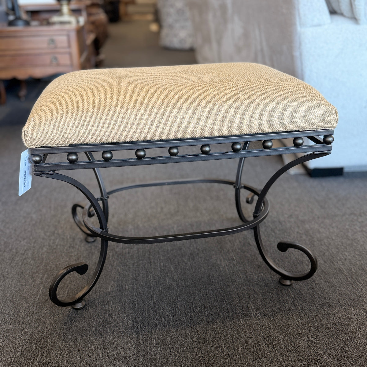 A wrought-iron vanity bench featuring scrollwork legs, studded metal apron trim, and a textured beige tweed upholstered cushion in excellent condition.