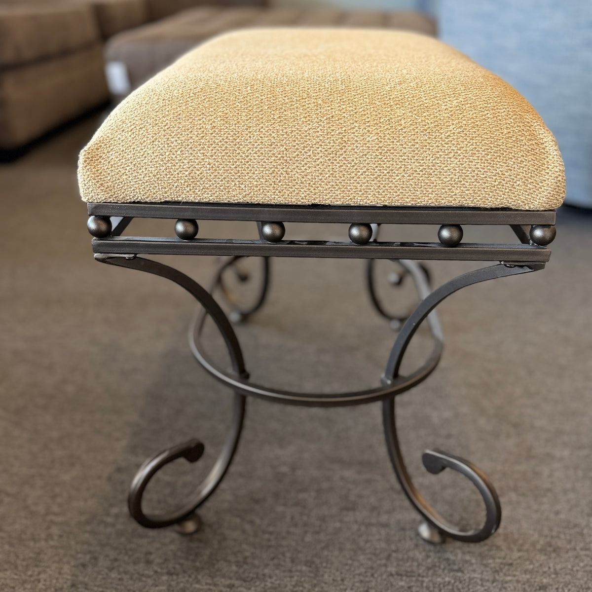 A wrought-iron vanity bench featuring scrollwork legs, studded metal apron trim, and a textured beige tweed upholstered cushion in excellent condition.