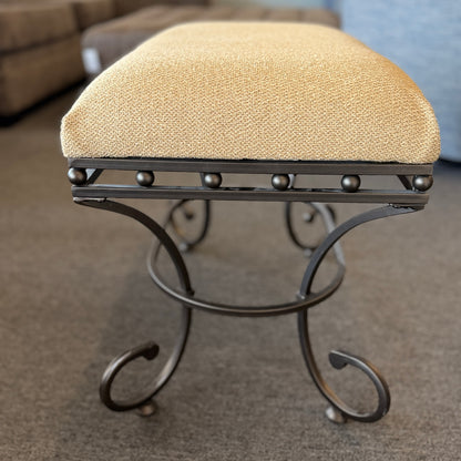 A wrought-iron vanity bench featuring scrollwork legs, studded metal apron trim, and a textured beige tweed upholstered cushion in excellent condition.